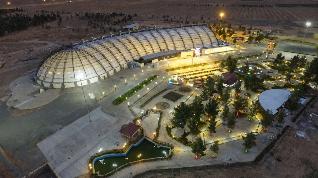An aerial view at dusk of the domed Abotab water park complex at Ghadeer Park, its vast curved glass roof lit up beside landscaped gardens and ponds