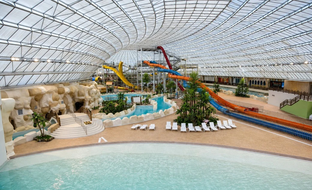The interior of the Abotab water park, a huge hall under a vaulted glass roof with tangled blue and orange water slides, a wave pool and sun loungers