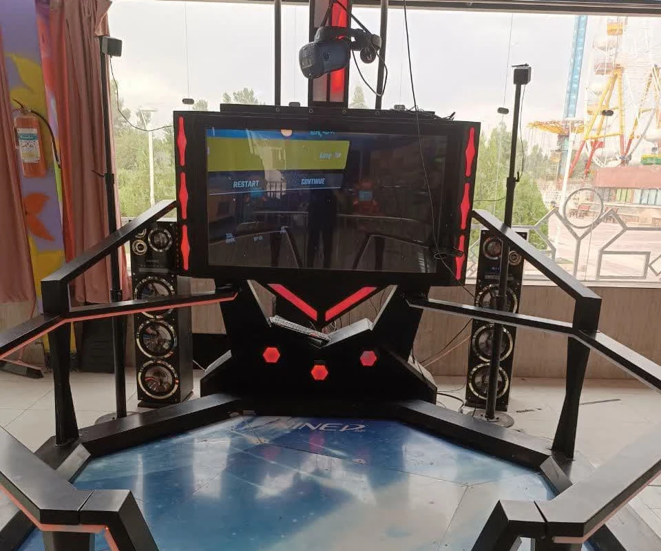 A virtual-reality standing simulator with a large screen and speakers inside an indoor amusement park in Qum