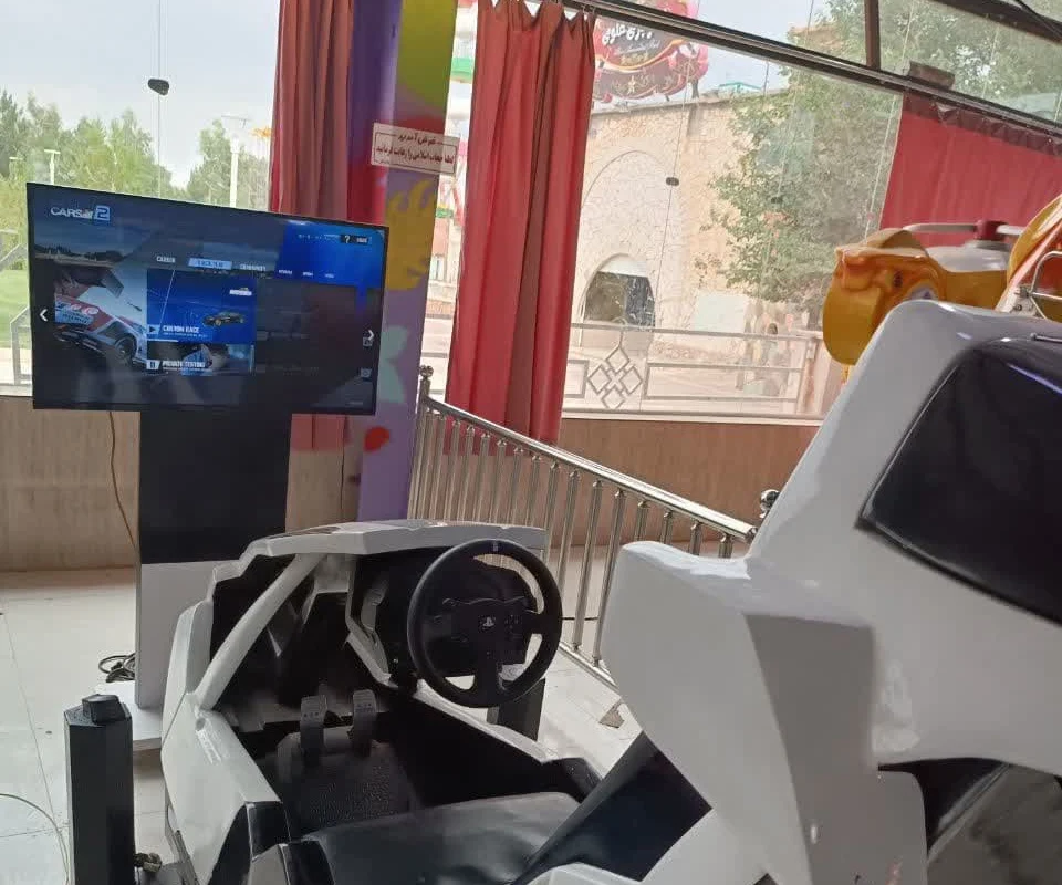 A white racing-simulator cockpit with steering wheel and pedals facing a screen showing a driving game