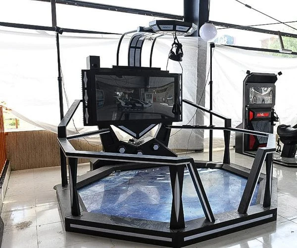 A black virtual-reality platform rig with an overhead harness and screen, arcade cabinets behind