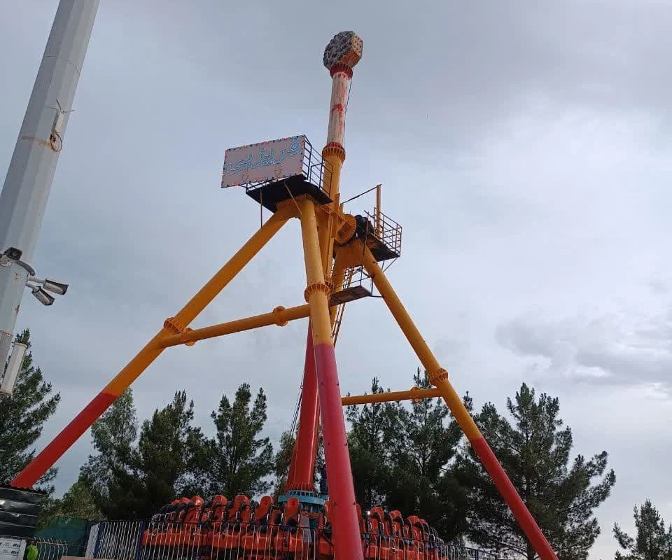 A red and yellow Frisbee pendulum ride towering over an outdoor amusement park in Qum, its arm raised against a cloudy sky