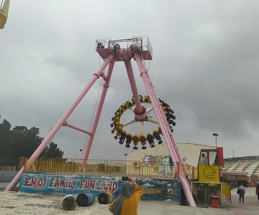 A large pink pendulum ride swinging a circular gondola of seated riders high into a cloudy sky at an outdoor amusement park in Qum