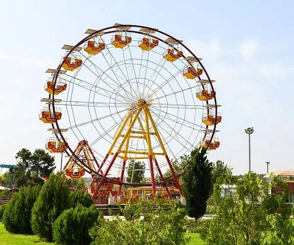 A red and yellow ferris wheel turning above conifers and lawns at Alavi Park