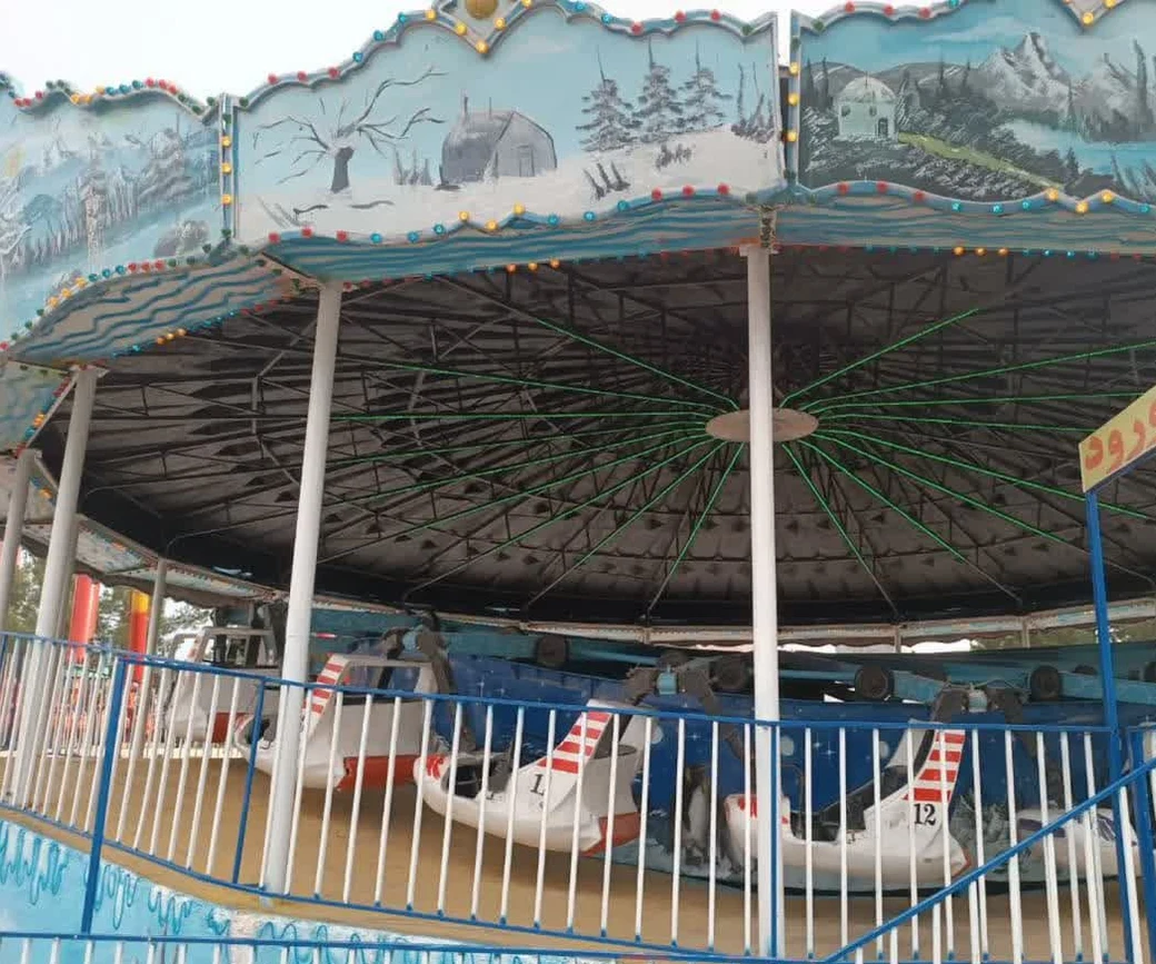 A hand-painted blue carousel with boat-shaped cars and coloured bulbs at an outdoor amusement park in Qum
