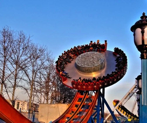A spinning tilt-disc ride carrying riders around at dusk, with roller-coaster track in the foreground