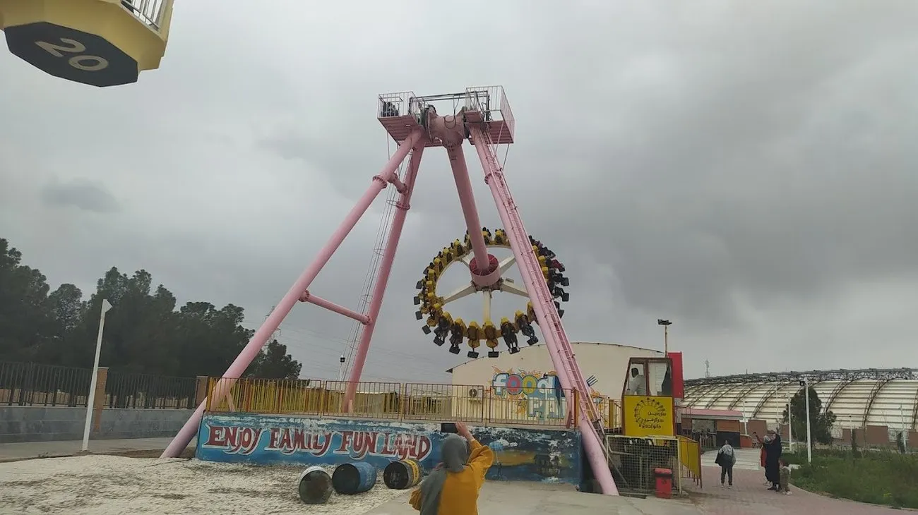 A large pink pendulum ride swinging a circular gondola of seated riders high into a cloudy sky at an outdoor amusement park in Qum