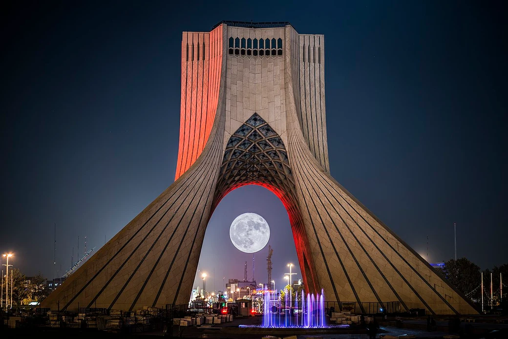 The Azadi Tower in Tehran at dusk, its white marble arch framing a full moon, with a fountain playing at its base