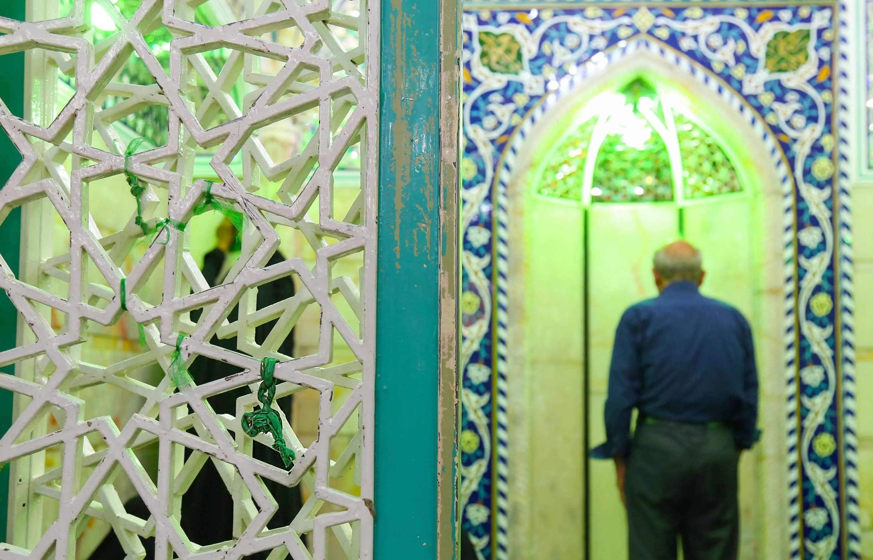 The green-lit mihrab inside Bayt al-Nur in Qum, framed by a white lattice screen, with a pilgrim standing in prayer before its blue-tiled arch