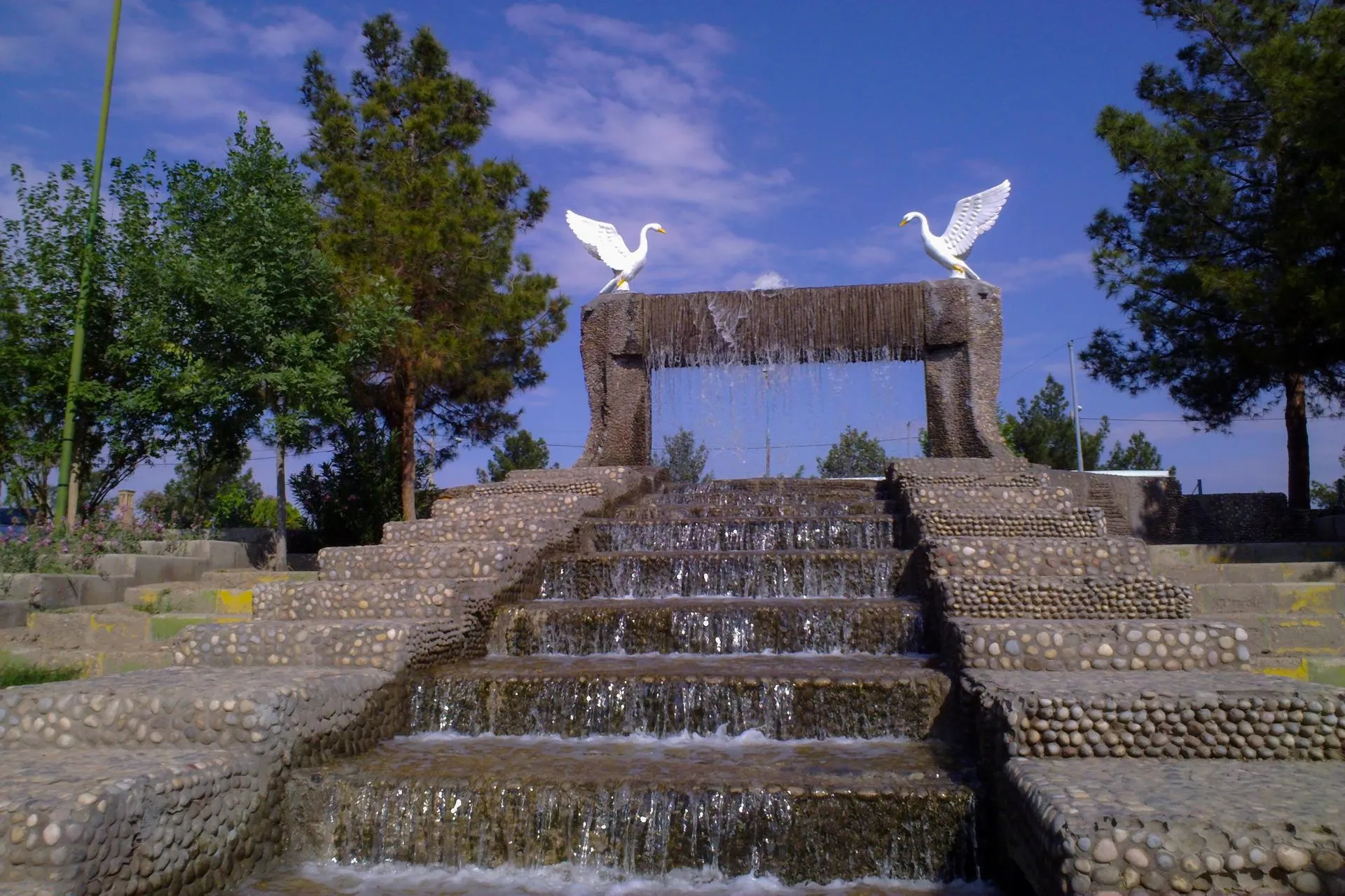 The stepped stone fountain at Bonyadi Park, water cascading down pebble-clad steps beneath two white swan sculptures on a sunny day