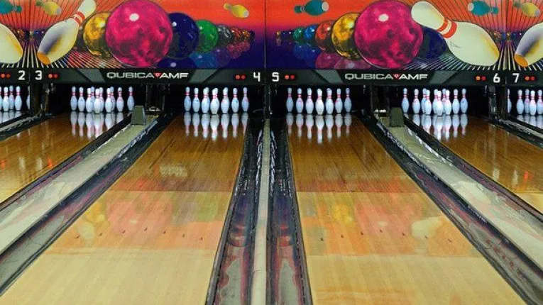 A row of polished ten-pin bowling lanes with pins set and a colourful mural of bowling balls above the QubicaAMF scoreboards