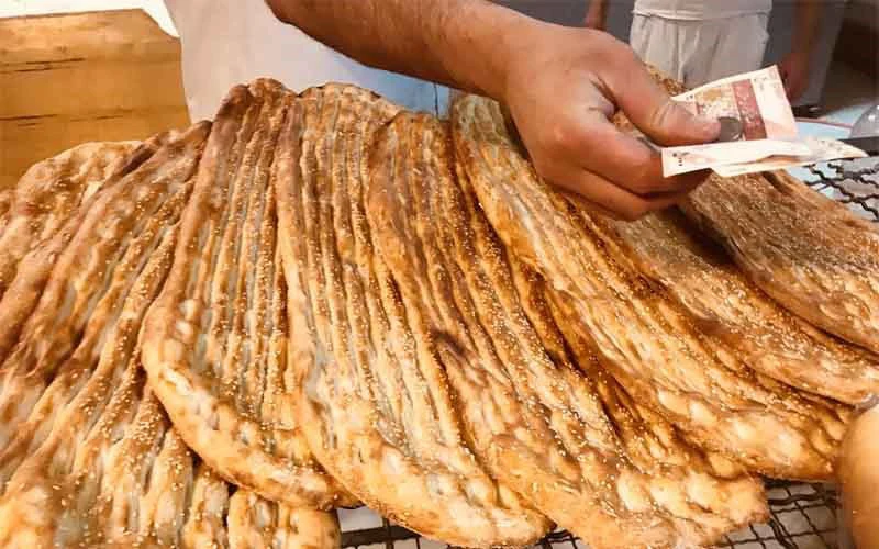 Long, ridged, sesame-topped Barbari loaves stacked at a bakery, with a customer handing over cash