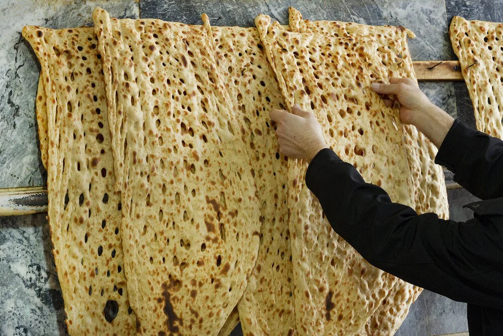 Long, triangular Sangak flatbreads with a bubbled, pebble-baked surface hanging on a rack, one being lifted by hand