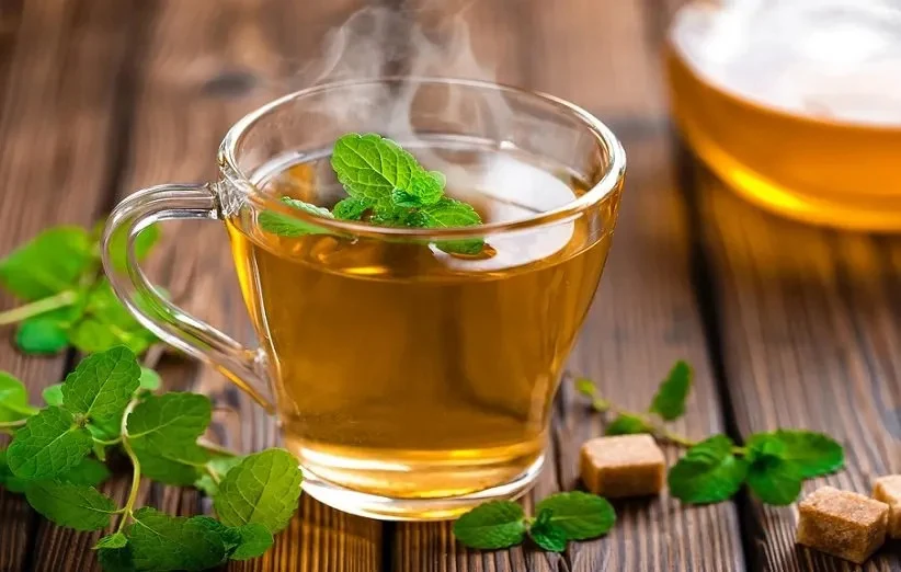 A steaming glass cup of pale herbal tea garnished with fresh mint leaves, with mint sprigs and brown sugar cubes on the table beside it