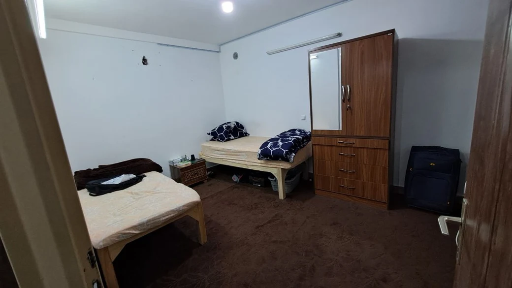 A shared SIOS dormitory bedroom with two single beds, a wooden wardrobe and chest of drawers, and a suitcase against the wall
