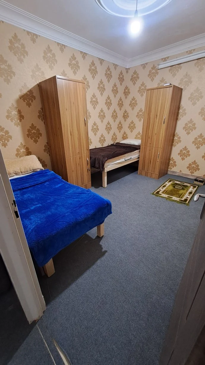 Another SIOS dormitory bedroom with two single beds, tall wooden wardrobes, patterned wallpaper and a small prayer mat on the floor