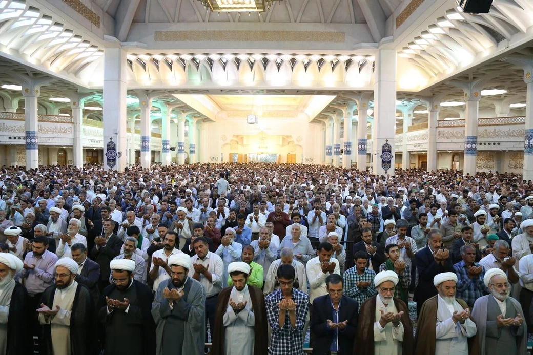 A vast congregation of men, many in clerical dress, standing with hands raised in supplication during Eid prayers inside a large mosque in Qum