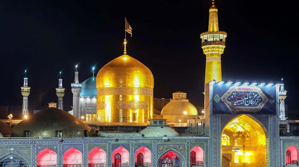 The shrine of Imam al-Ridha (a) in Mashhad at night, its golden dome and minaret floodlit above the tiled courtyard arcades, with a black mourning banner hung across the iwan