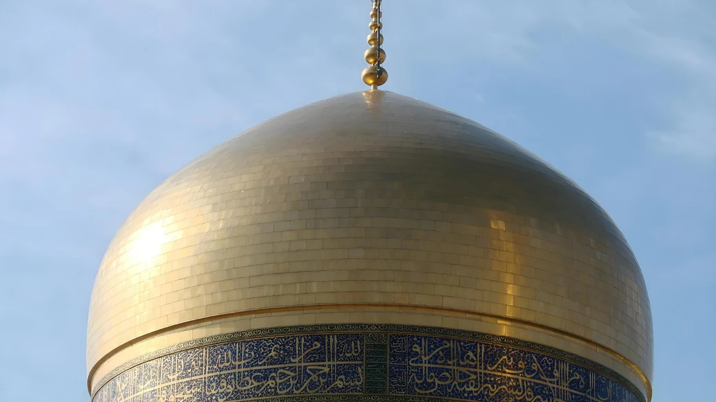 The golden dome of the shrine of Imam al-Ridha (a) in daylight, its gilded tiles topped by a finial and banded with blue-and-gold calligraphic tilework