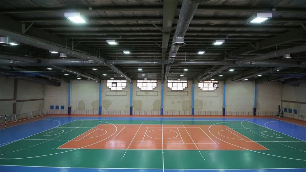 A large indoor sports hall in Qum with a green and orange futsal court marked out for multiple sports, ringed by low barriers under an exposed-ductwork ceiling
