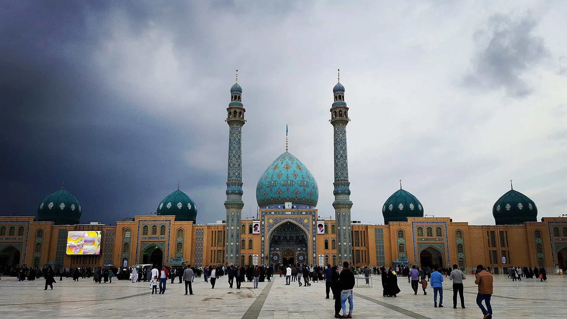 The main façade of Jamkaran Mosque in daylight — a turquoise tiled central dome flanked by twin tiled minarets and four smaller green domes, with pilgrims crossing the wide marble courtyard beneath a storm-dark sky