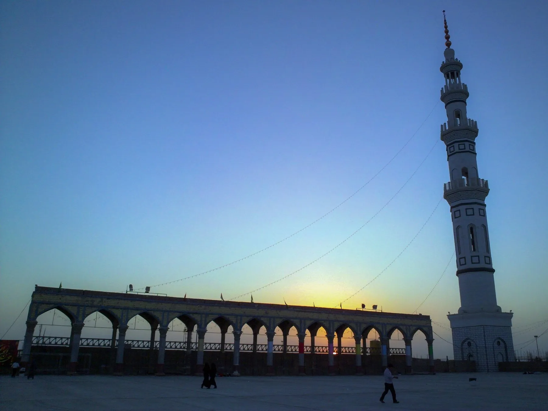 Jamkaran Mosque on the edge of Qum at dusk, its tall white minaret and arcaded courtyard wall silhouetted against a glowing sky