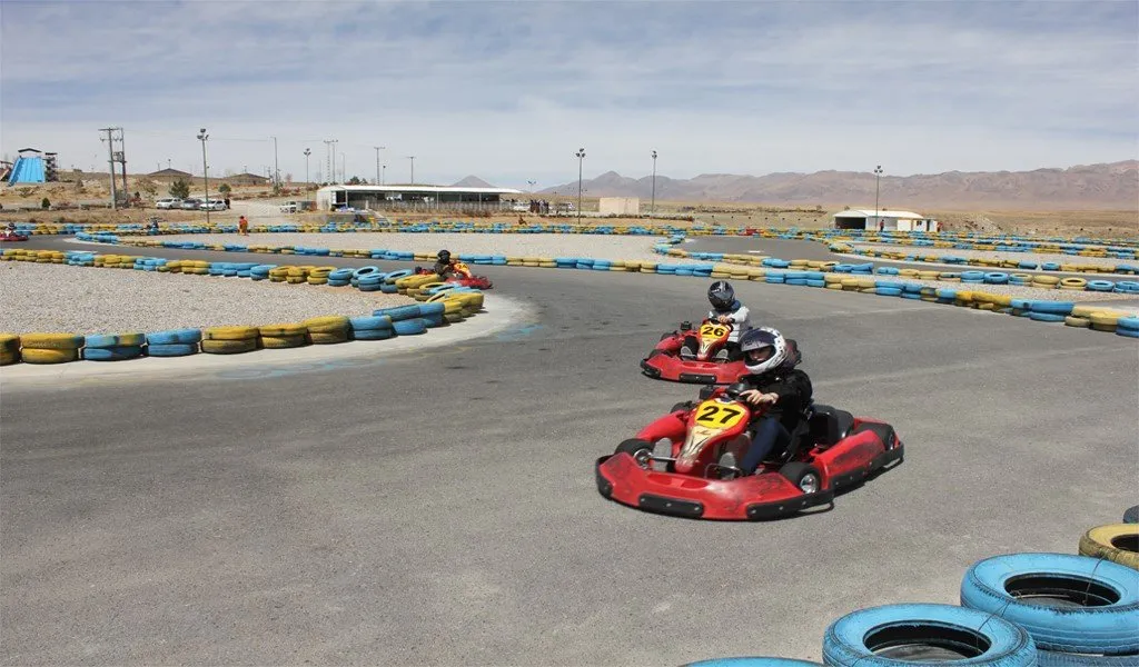 Two go-karts numbered 26 and 27 racing around a tarmac track lined with blue and yellow tyres, with desert hills in the background near Qum