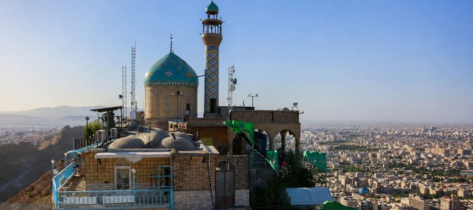 The shrine atop Khizr Mountain, its turquoise dome and patterned minaret rising above a rocky summit terrace lined with blue railings and green flags, overlooking the dense sprawl of Qum below
