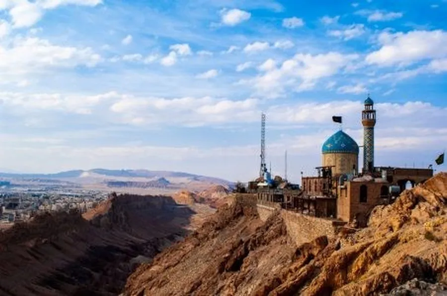 A small blue-domed mosque perched on the rocky summit of Khizr Mountain, overlooking the desert plain around Qum under a bright sky