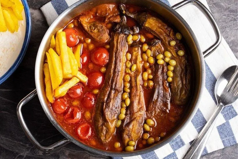 A pot of Khoresht Bademjan — whole roasted aubergines simmered in a red tomato sauce with cherry tomatoes, chickpeas and unripe grapes — served with a plate of saffron rice and fries