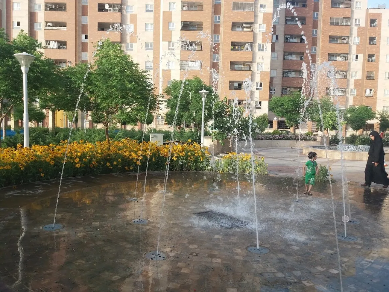 The water play area at Koodak Park in Qum, where a child plays among arcing jets of water on a paved splash pad, with flowerbeds and apartment blocks behind