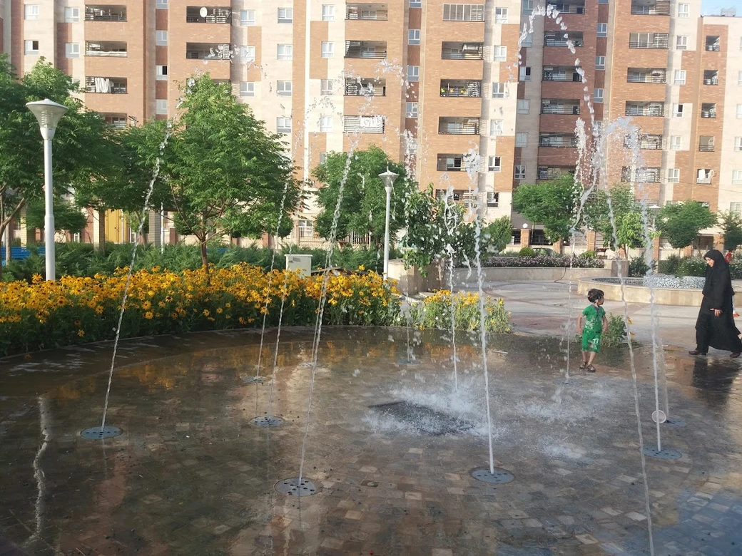 The water play area at Koodak Park in Qum, where a child plays among arcing jets of water on a paved splash pad, with flowerbeds and apartment blocks behind