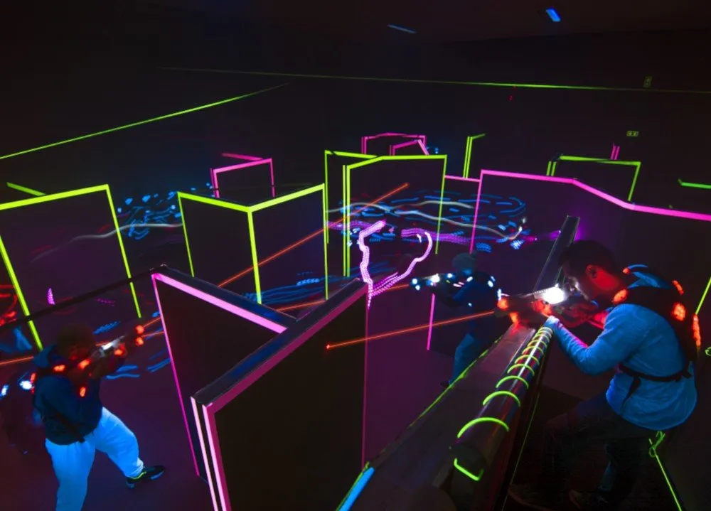 Players in a dark laser-tag arena lit by neon green and pink lines, aiming infrared light guns from behind glowing barriers