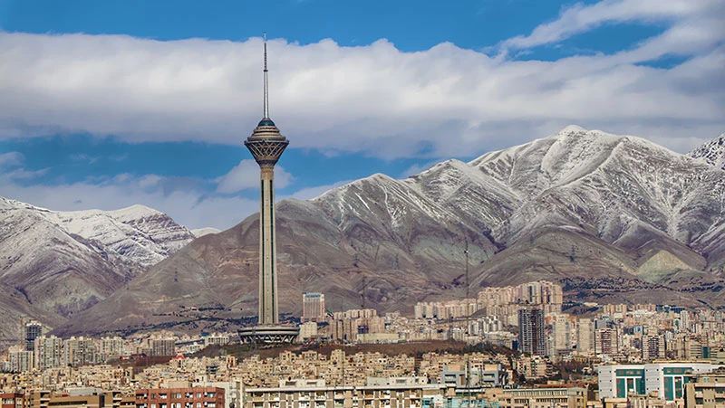 The Milad Tower rising above the Tehran skyline, its slender concrete shaft topped by a lattice-clad observation pod, with the snow-dusted Alborz mountains behind