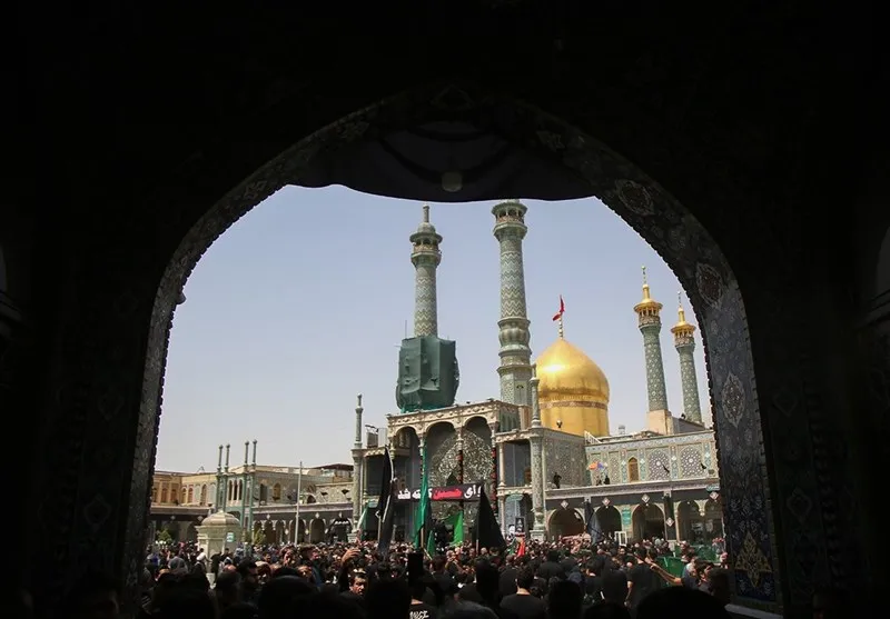 A large crowd of mourners gathered in the courtyard of the Lady Masumah (a) shrine in Qum, draped in black with mourning flags, viewed through a tiled archway with the golden dome behind