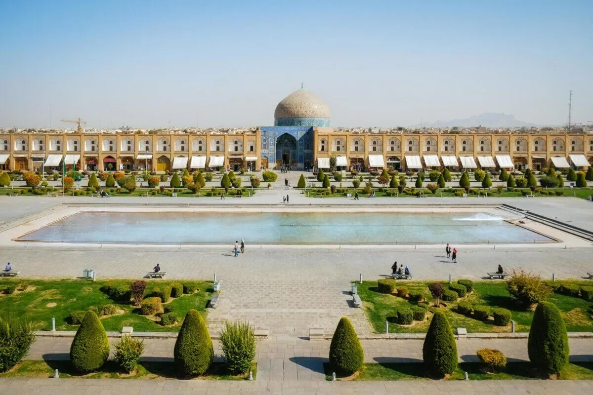 Naqsh-e Jahan Square in Isfahan, a vast rectangular public square with a long central pool and manicured gardens, the tiled dome of the Sheikh Lutfullah Mosque rising behind arcaded shopfronts