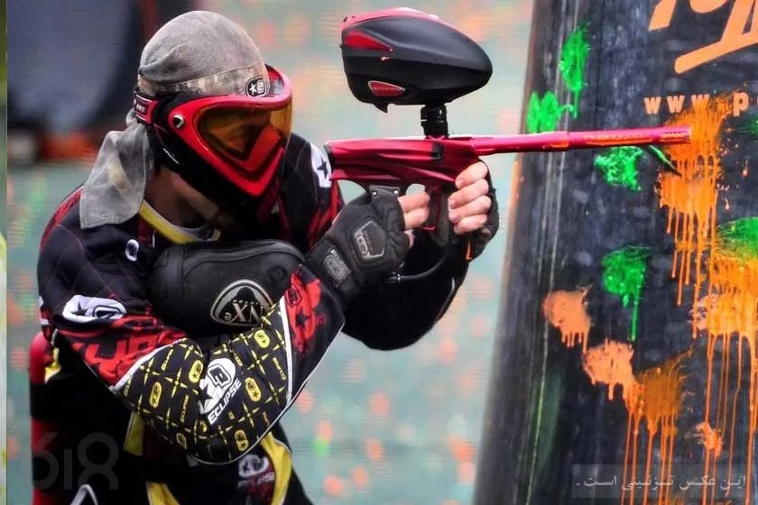 A masked paintball player in full protective gear taking aim with a red marker from behind cover, orange and green paint splattered on the barrier beside him