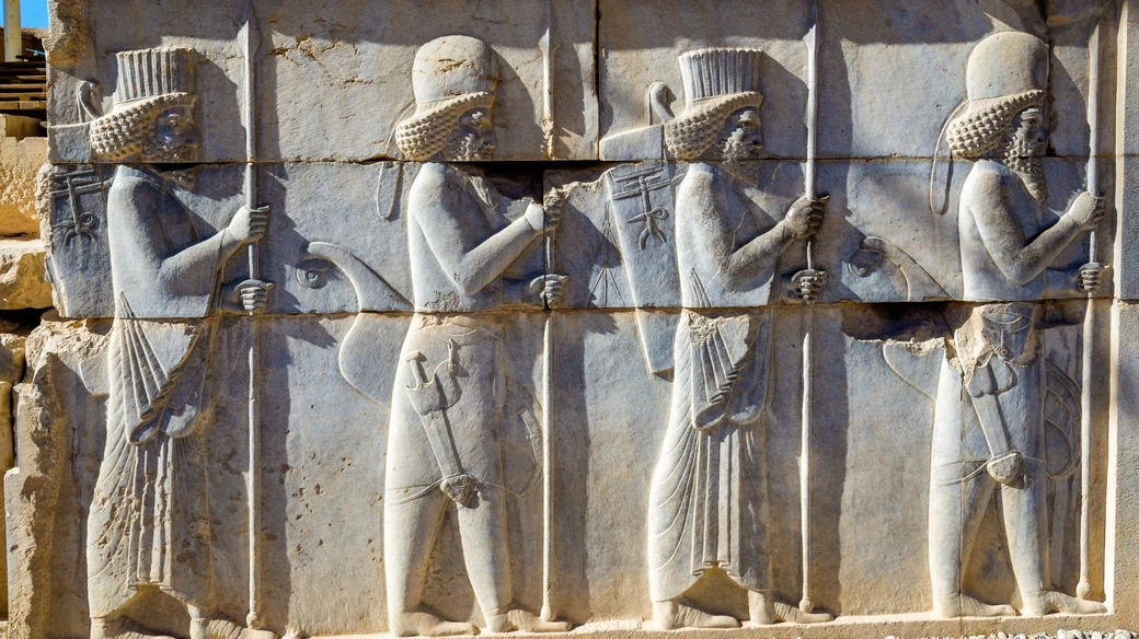 A carved stone relief at Persepolis showing four Persian and Median guards in procession, each carrying a spear, bow and quiver, beneath a rosette border