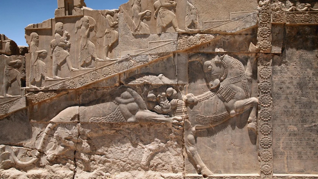 A relief on the Apadana staircase at Persepolis showing a lion attacking a bull, with a tribute procession and a cuneiform inscription panel alongside