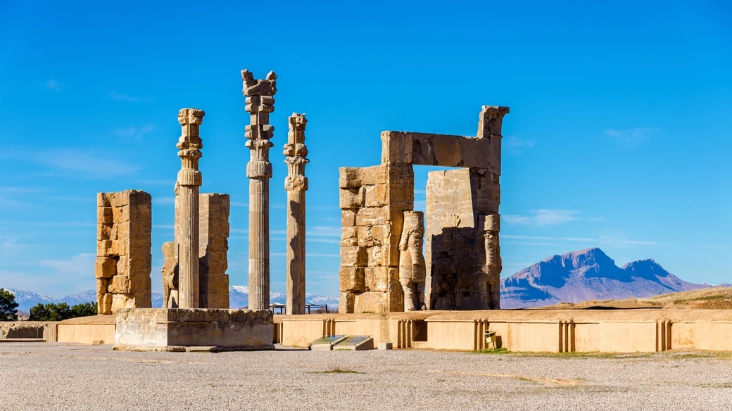 The Gate of All Nations at Persepolis, its standing columns and stone doorways silhouetted against a clear blue sky with the Zagros mountains behind