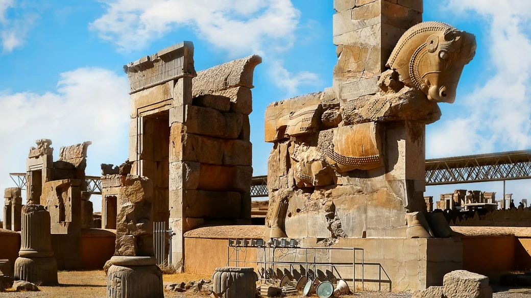 The Hall of a Hundred Columns at Persepolis, with a carved double horse-head capital resting on a ruined pillar and the Tachara palace beyond