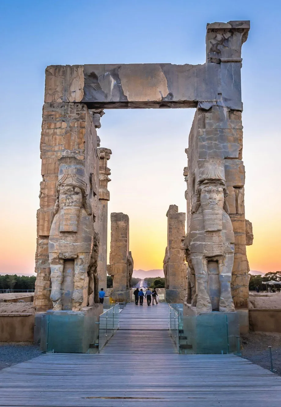 The Gate of All Nations at Persepolis at sunset, two towering stone gateways flanked by carved lamassu figures, with visitors walking along a boardwalk between them
