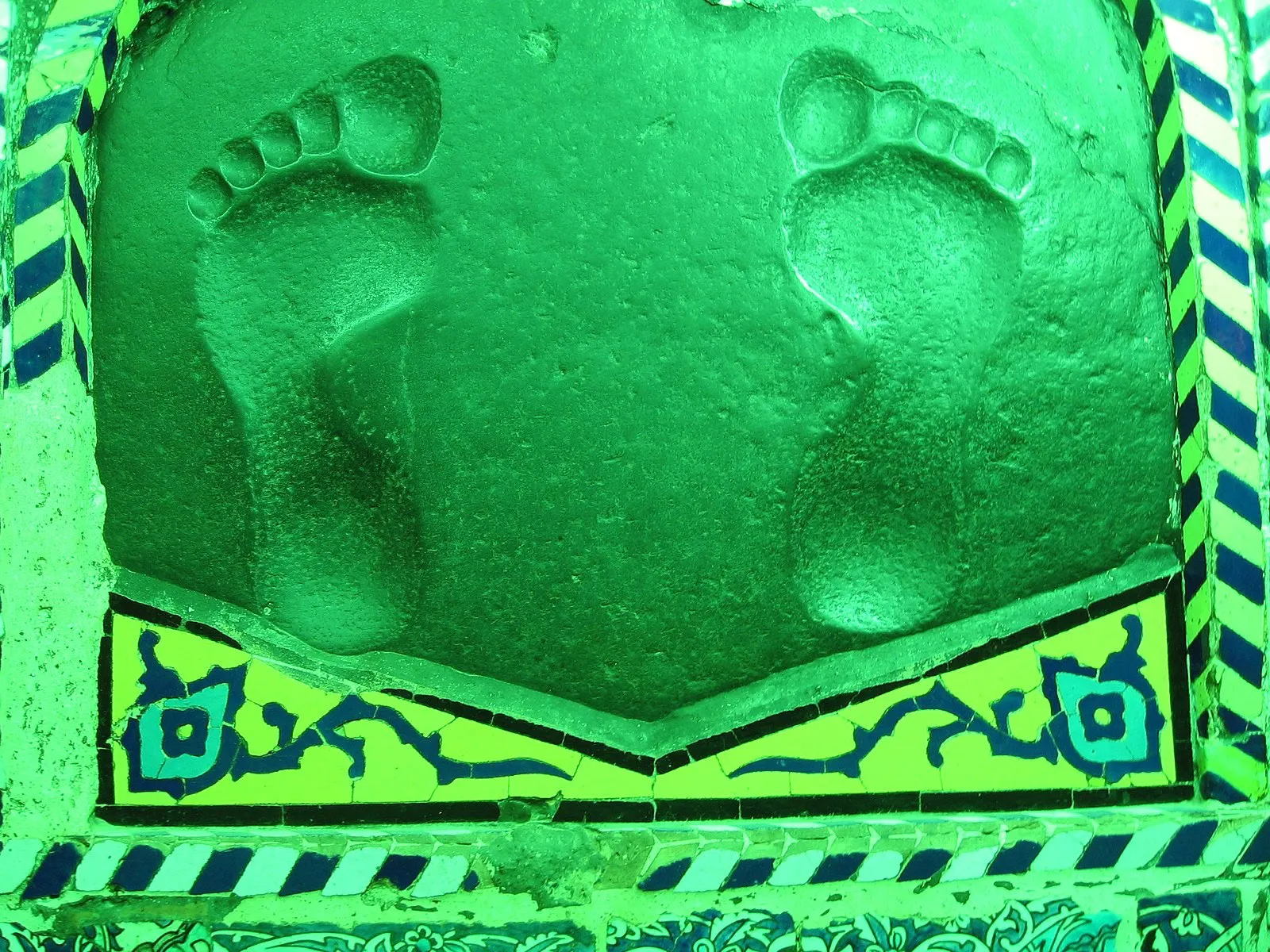 Two footprints impressed into a stone at the Qadam Gah near Neyshabur, lit with green light and framed by turquoise and blue tilework