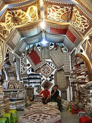 A carpet shop inside a covered Iranian bazaar, its domed ceiling and walls hung with patterned carpets and kilims, with two shopkeepers seated among stacks of rugs