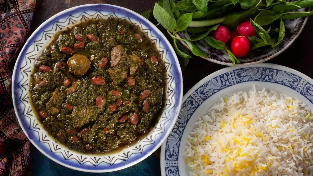 A bowl of Qorma Sabzi — a dark green herb stew with red kidney beans and chunks of lamb — served with a plate of saffron-flecked rice and a side of fresh herbs and radishes