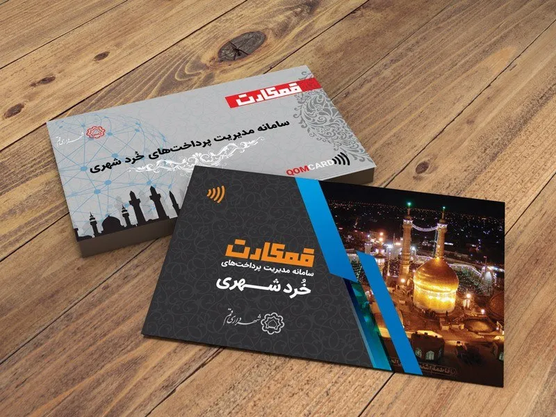 Two Qum Card contactless smartcards resting on a wooden surface, one grey and one black, both showing the QOM CARD logo and a contactless symbol