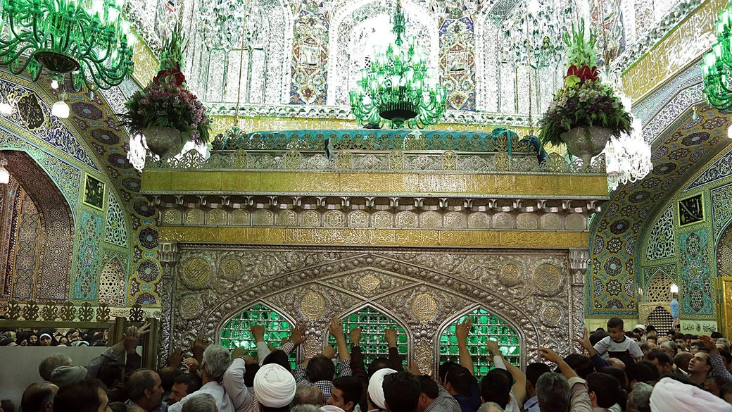 The interior of the shrine of Imam al-Ridha (a) in Mashhad, with crowds of pilgrims reaching towards the silver-and-gold zarih beneath dazzling mirrorwork walls and green chandeliers