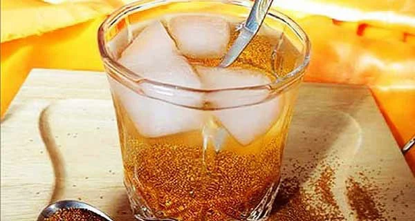 A glass of Sharbat-e Khakshir — swollen flixweed seeds in a golden, iced drink — with a spoon and scattered seeds beside it