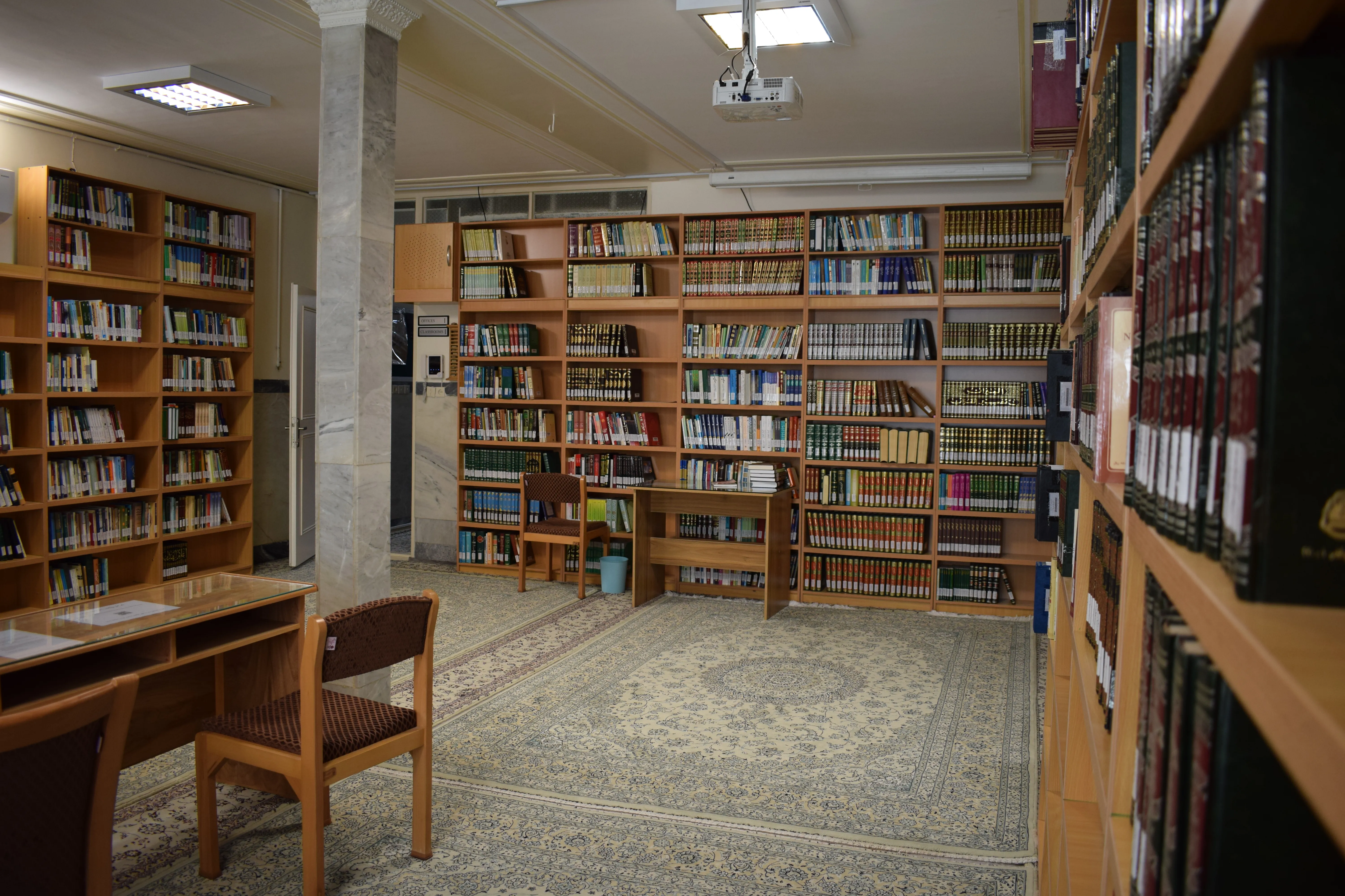 Wider view of the SIOS library study area, showing shelves of coloured book spines, individual study desks and chairs on a patterned Persian carpet