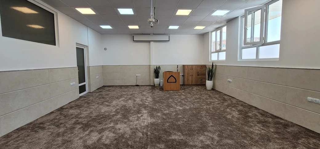 The SIOS prayer hall: a spacious carpeted room with a small wooden lectern bearing a mihrab-shaped emblem, and large windows letting in daylight